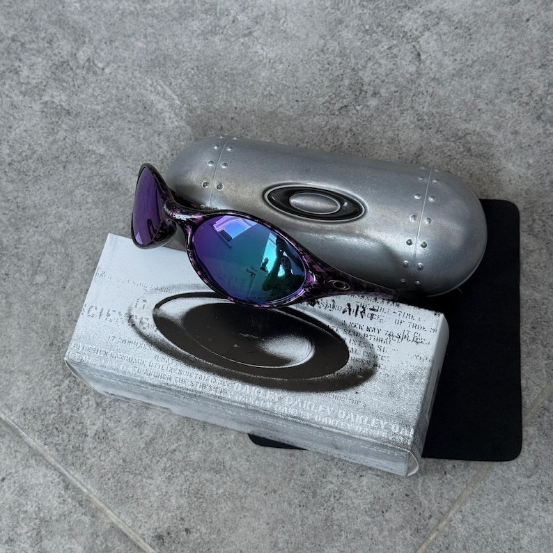 Puede incluir: Gafas de sol con montura moteada en morado y negro y lentes iridiscentes. Se muestran con un estuche plateado y una caja con texto. Las lentes reflejan tonos azules y verdes.