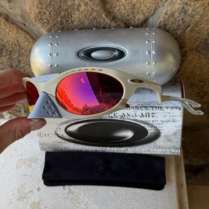 May include: Off-white Oakley sunglasses with red mirrored lenses, a gray nose bridge, and a silver case. The sunglasses are displayed with a black case and a box with the Oakley logo.