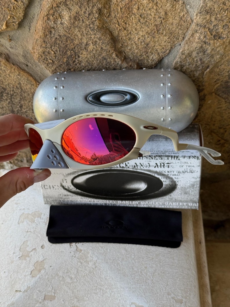 May include: Off-white Oakley sunglasses with red mirrored lenses, a gray nose bridge, and a silver case. The sunglasses are displayed with a black case and a box with the Oakley logo.