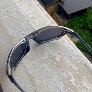 May include: Sleek, modern sunglasses with a silver and black frame. The lenses are dark, providing shade. The design is sporty and streamlined, suitable for outdoor activities. The sunglasses are resting on a weathered, light-colored surface.