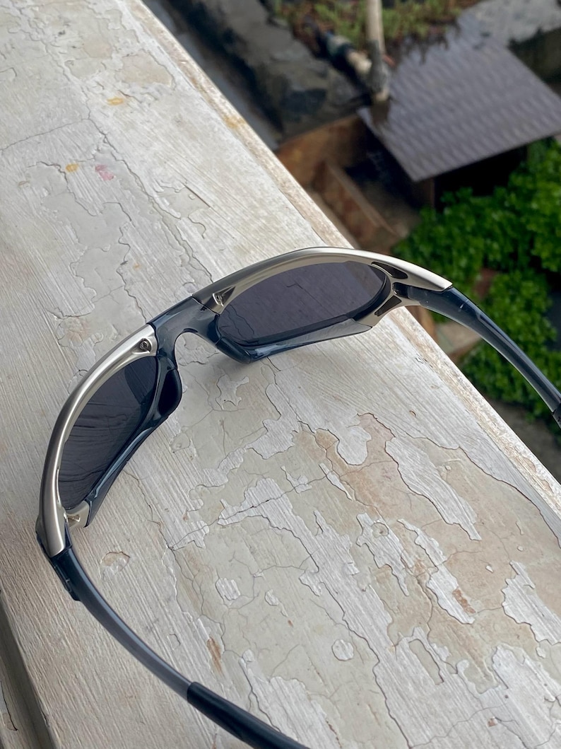 May include: Sleek, modern sunglasses with a silver and black frame. The lenses are dark, providing shade. The design is sporty and streamlined, suitable for outdoor activities. The sunglasses are resting on a weathered, light-colored surface.
