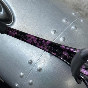 Puede incluir: Primer plano de unas gafas de sol con estampado negro y morado. La montura es negra con un dise&ntilde;o floral morado. El fondo es una superficie met&aacute;lica plateada con remaches.