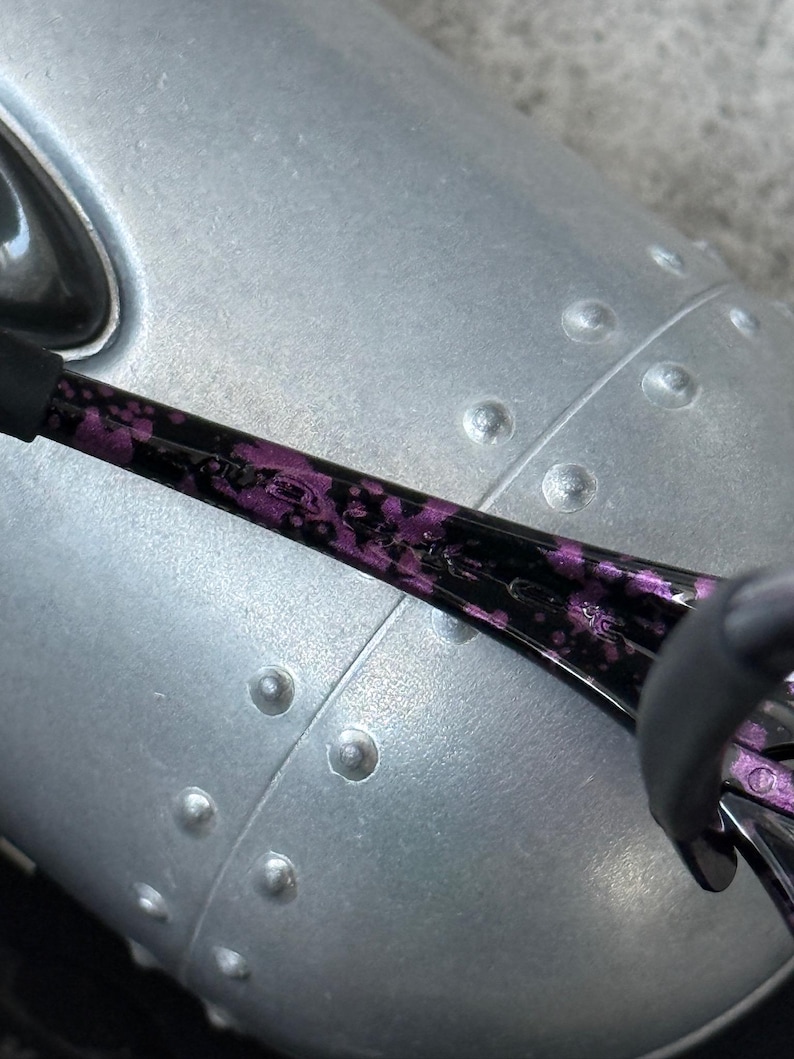 Puede incluir: Primer plano de unas gafas de sol con estampado negro y morado. La montura es negra con un dise&ntilde;o floral morado. El fondo es una superficie met&aacute;lica plateada con remaches.