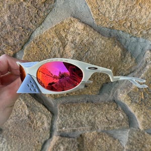 May include: Off-white sports sunglasses with red mirrored lenses. The sunglasses have a unique design with a light blue nose bridge and temples. The lenses reflect the surrounding environment. The sunglasses are set against a stone background.