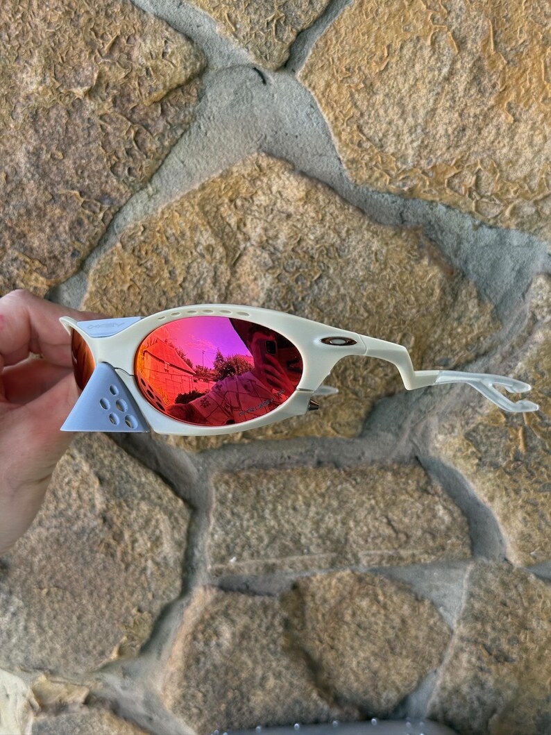 May include: Off-white sports sunglasses with red mirrored lenses. The sunglasses have a unique design with a light blue nose bridge and temples. The lenses reflect the surrounding environment. The sunglasses are set against a stone background.