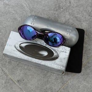 Puede incluir: Gafas de sol Oakley moradas y negras con lentes azules reflectantes, sobre un estuche plateado y una caja con el logotipo de Oakley. La caja tiene texto. Las gafas de sol son de estilo deportivo.