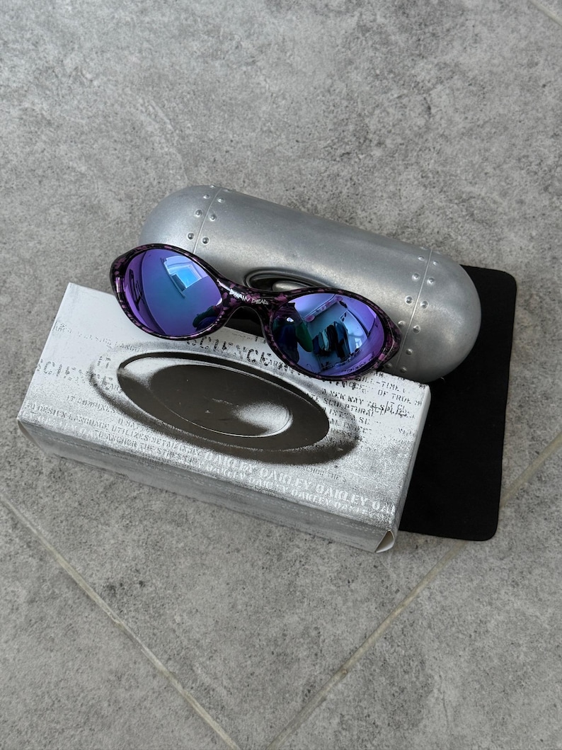 Puede incluir: Gafas de sol Oakley moradas y negras con lentes azules reflectantes, sobre un estuche plateado y una caja con el logotipo de Oakley. La caja tiene texto. Las gafas de sol son de estilo deportivo.
