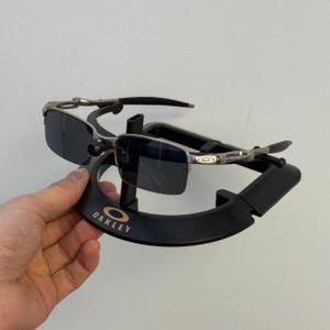 May include: Oakley sunglasses with dark lenses on a black display stand. The stand features the Oakley logo in gold lettering. The sunglasses have a silver frame and black arms.