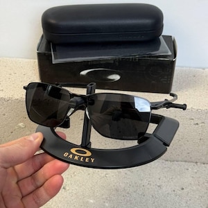 May include: Black Oakley sunglasses with a black case and box. The sunglasses are displayed on a black stand with the Oakley logo in gold. The lenses are dark, and the frames are black.