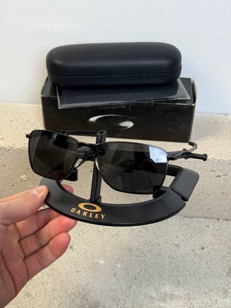 May include: Black Oakley sunglasses with a black case and box. The sunglasses are displayed on a black stand with the Oakley logo in gold. The lenses are dark, and the frames are black.