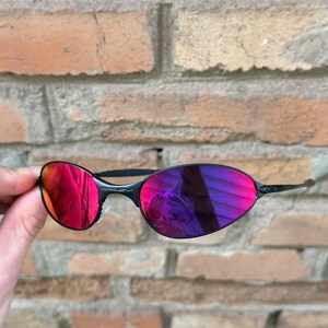 May include: Black rimless sunglasses with vivid red and purple lenses. The lenses reflect the surroundings, creating a dynamic visual effect. The sunglasses have a sleek, modern design.