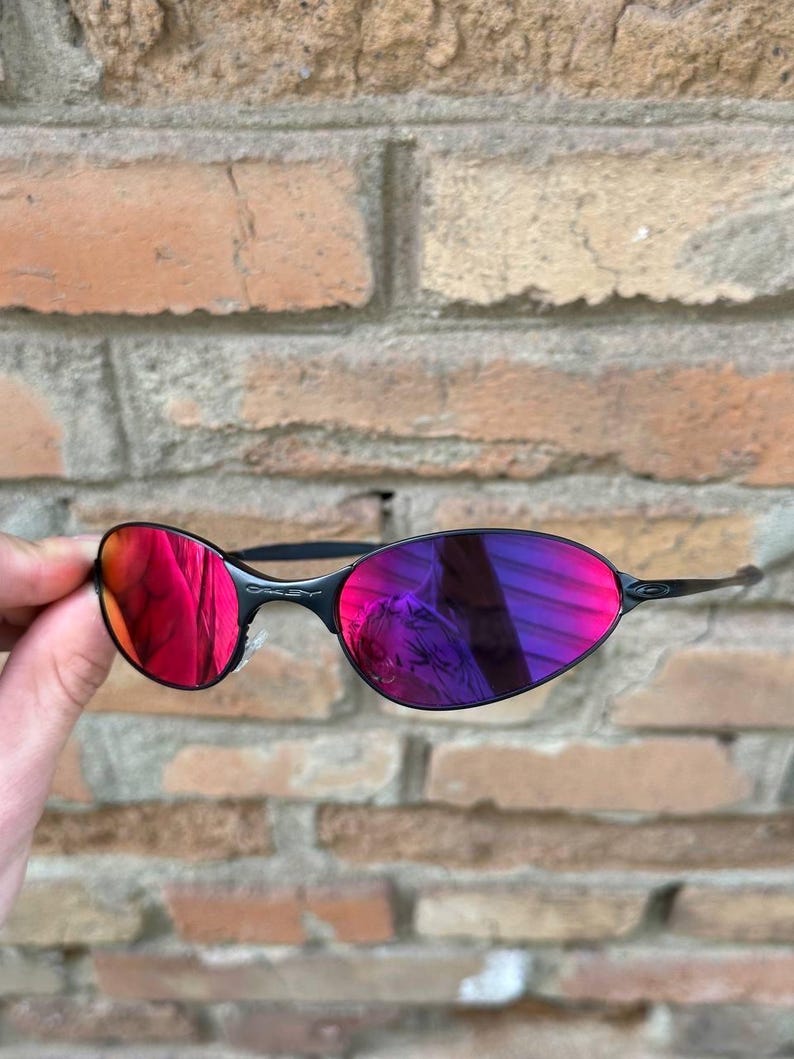 May include: Black rimless sunglasses with vivid red and purple lenses. The lenses reflect the surroundings, creating a dynamic visual effect. The sunglasses have a sleek, modern design.