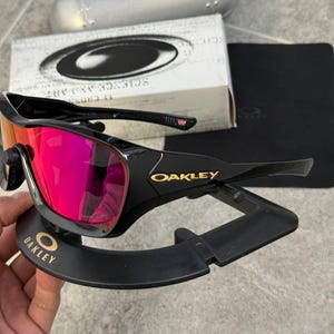 May include: Black Oakley sunglasses with red mirrored lenses, displayed on a black stand. The Oakley logo is visible in gold on the frame and stand. A silver case and a box with the Oakley logo are in the background.