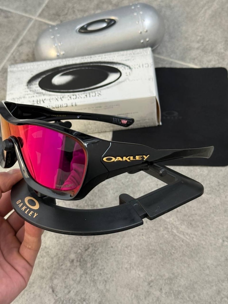 May include: Black Oakley sunglasses with red mirrored lenses, displayed on a black stand. The Oakley logo is visible in gold on the frame and stand. A silver case and a box with the Oakley logo are in the background.
