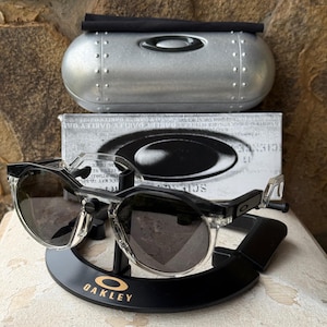 May include: Clear-framed sunglasses with dark lenses rest on a black display stand with the Oakley logo in gold. A silver case and a box with the Oakley logo are behind the sunglasses. The sunglasses have a modern, sporty design.