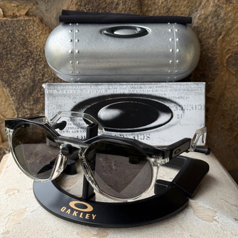 May include: Clear-framed sunglasses with dark lenses rest on a black display stand with the Oakley logo in gold. A silver case and a box with the Oakley logo are behind the sunglasses. The sunglasses have a modern, sporty design.