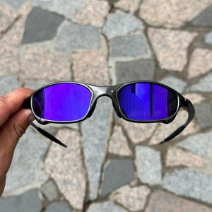 May include: Black sunglasses with purple mirrored lenses. The sunglasses have a sleek, modern design with a dark frame and curved temples. The lenses reflect a vibrant purple hue, offering a stylish and functional accessory.