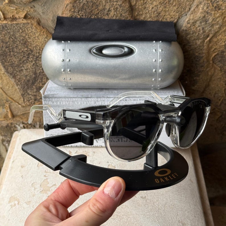 May include: Clear-framed sunglasses with dark lenses rest on a black display stand. The stand has the brand name "Oakley" in gold lettering. A silver case with the Oakley logo and a black cloth are in the background.