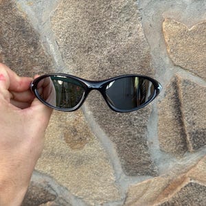 May include: Black sunglasses with a curved frame and mirrored lenses. The sunglasses have a small white accent on the outer corners. The image is of the sunglasses held in front of a stone wall.