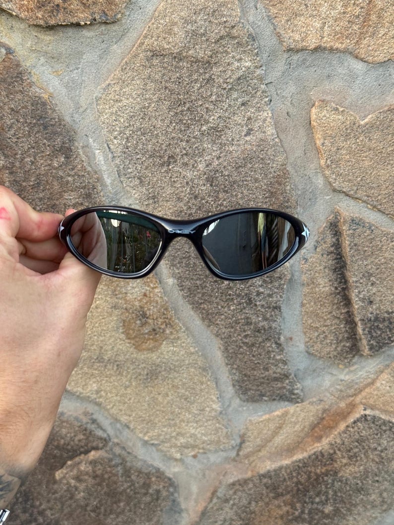 May include: Black sunglasses with a curved frame and mirrored lenses. The sunglasses have a small white accent on the outer corners. The image is of the sunglasses held in front of a stone wall.
