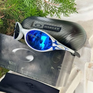 May include: White and blue Oakley sunglasses with reflective blue lenses. The sunglasses have a unique design with a white frame and blue accents. A black case with the Oakley logo and a black soft case are also included.