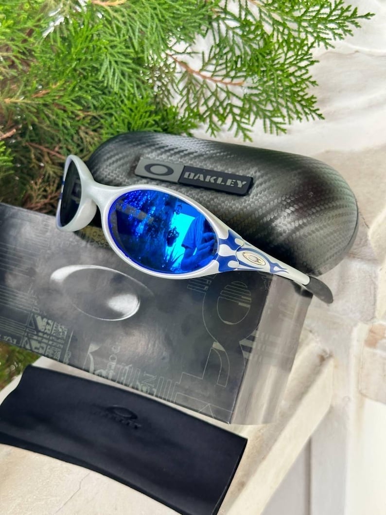 May include: White and blue Oakley sunglasses with reflective blue lenses. The sunglasses have a unique design with a white frame and blue accents. A black case with the Oakley logo and a black soft case are also included.