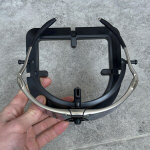 May include: A pair of sunglasses with a black frame and silver arms, held in a black display stand. The sunglasses have a curved design and dark lenses. The stand has a square shape with a central support.