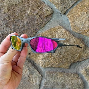 May include: Grey sunglasses with mirrored lenses reflecting yellow and pink hues. The sunglasses have a sleek, modern design with a grey frame and a curved shape. The lenses appear to be made of a reflective material.