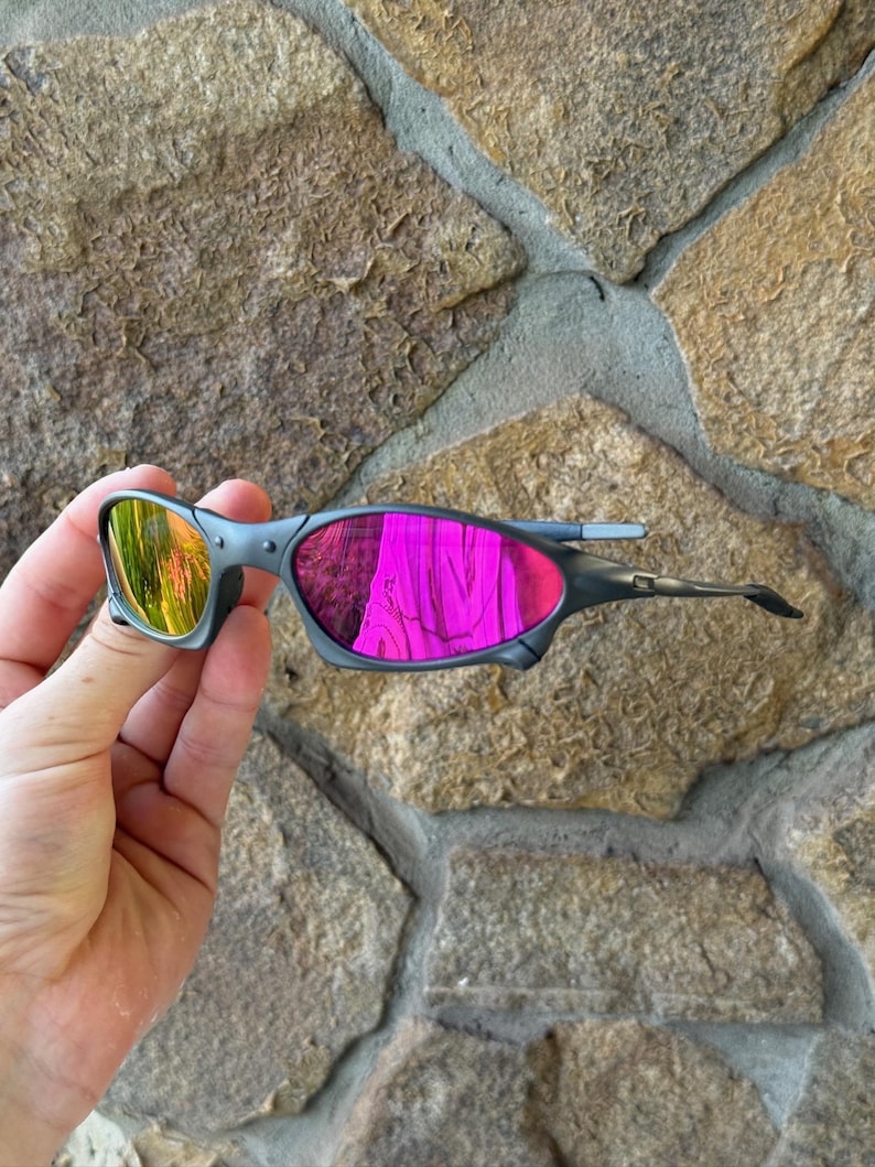May include: Grey sunglasses with mirrored lenses reflecting yellow and pink hues. The sunglasses have a sleek, modern design with a grey frame and a curved shape. The lenses appear to be made of a reflective material.
