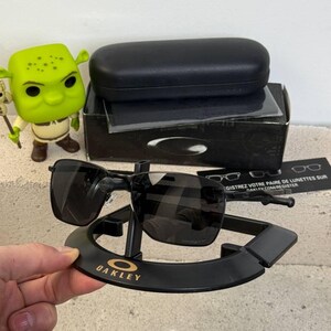 May include: Black Oakley sunglasses with a black case and display stand. The sunglasses have a rectangular frame and dark lenses. The stand has the Oakley logo in gold lettering. A Shrek figurine is visible in the background.