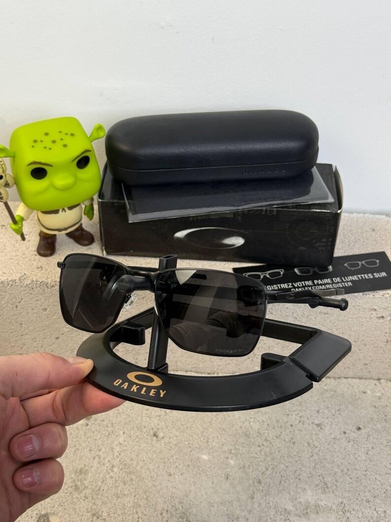 May include: Black Oakley sunglasses with a black case and display stand. The sunglasses have a rectangular frame and dark lenses. The stand has the Oakley logo in gold lettering. A Shrek figurine is visible in the background.