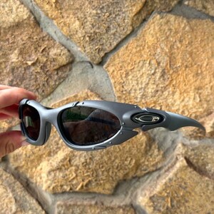 May include: Gray sunglasses with dark lenses. The sunglasses have a sporty, wraparound design with a curved frame and a small logo on the temple. The lenses are dark, and the frame has a matte finish.