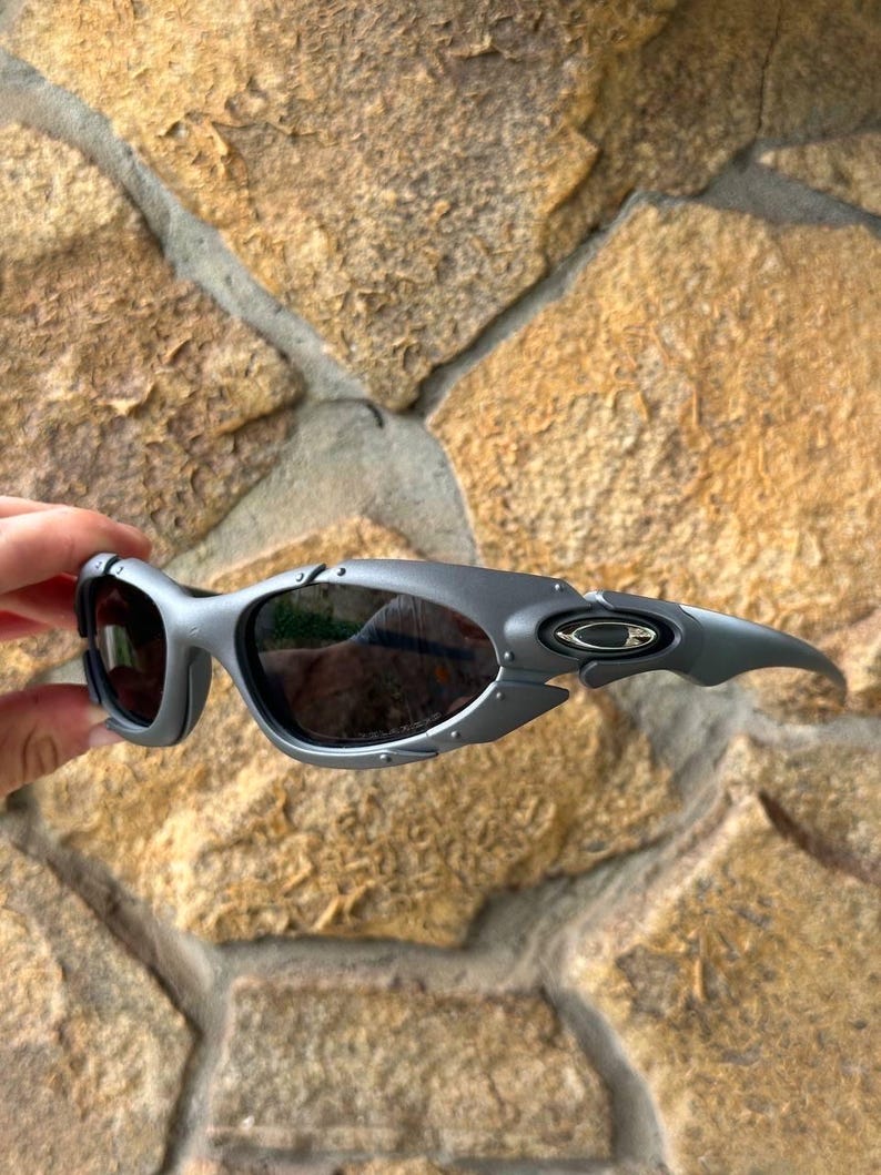 May include: Gray sunglasses with dark lenses. The sunglasses have a sporty, wraparound design with a curved frame and a small logo on the temple. The lenses are dark, and the frame has a matte finish.