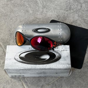 Op de afbeelding: Zwarte en rode Oakley zonnebril met een zilveren etui en een zwart zakje. De zonnebril heeft een zwart montuur met rode glazen en een vlammenontwerp aan de zijkant. Op de doos staat "SCIENCE" en "OAKLEY".