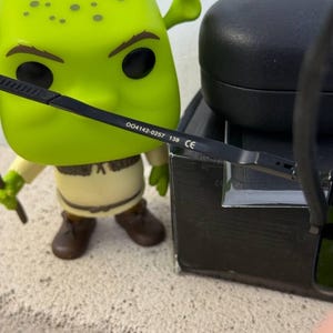 May include: A green Shrek Funko Pop figure with black eyeglasses. The eyeglasses have the text "004142-0257 139 CE" printed on the arm. A black glasses case is in the background.