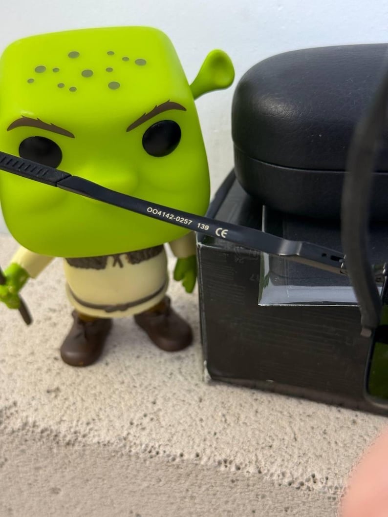 May include: A green Shrek Funko Pop figure with black eyeglasses. The eyeglasses have the text "004142-0257 139 CE" printed on the arm. A black glasses case is in the background.