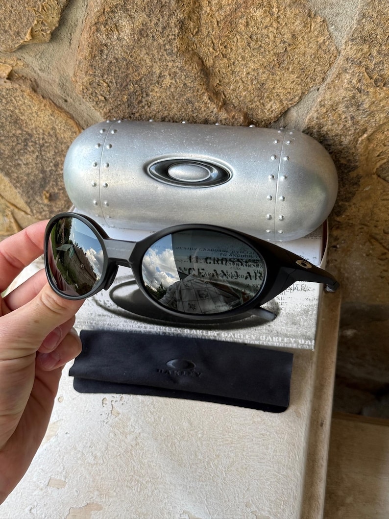 K&ouml;nnte beinhalten: Schwarze Oakley-Sonnenbrille mit silbernem Etui und schwarzem Mikrofasertuch. Die Sonnenbrille hat einen runden Rahmen und reflektierende Gl&auml;ser. Das Etui hat das Oakley-Logo und ein genietetes Design.
