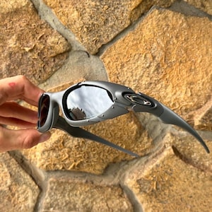 May include: Gray sunglasses with a curved design and dark lenses. The sunglasses have a sleek, modern aesthetic, with a metallic finish and a distinctive shape. The lenses reflect the surrounding environment.