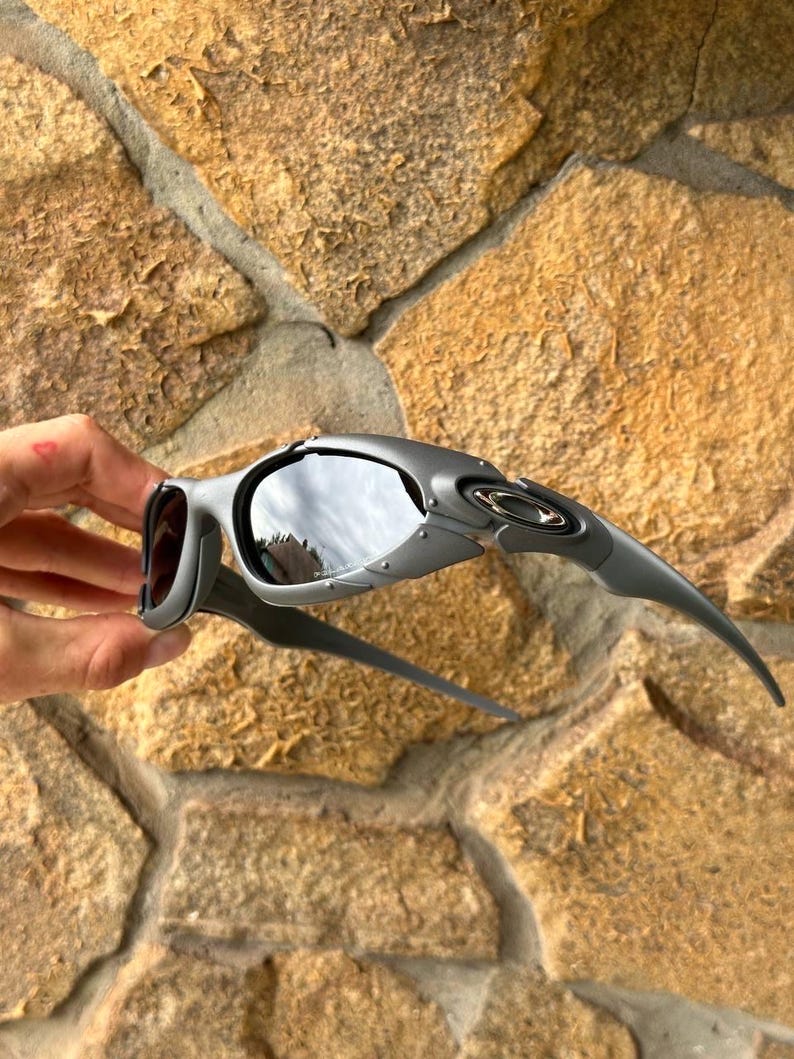 May include: Gray sunglasses with a curved design and dark lenses. The sunglasses have a sleek, modern aesthetic, with a metallic finish and a distinctive shape. The lenses reflect the surrounding environment.