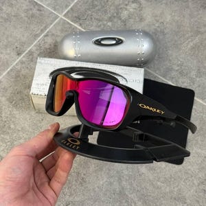 May include: Black Oakley sunglasses with a vibrant pink and orange lens. The sunglasses are displayed on a black stand with the Oakley logo. A silver case and a box are in the background.