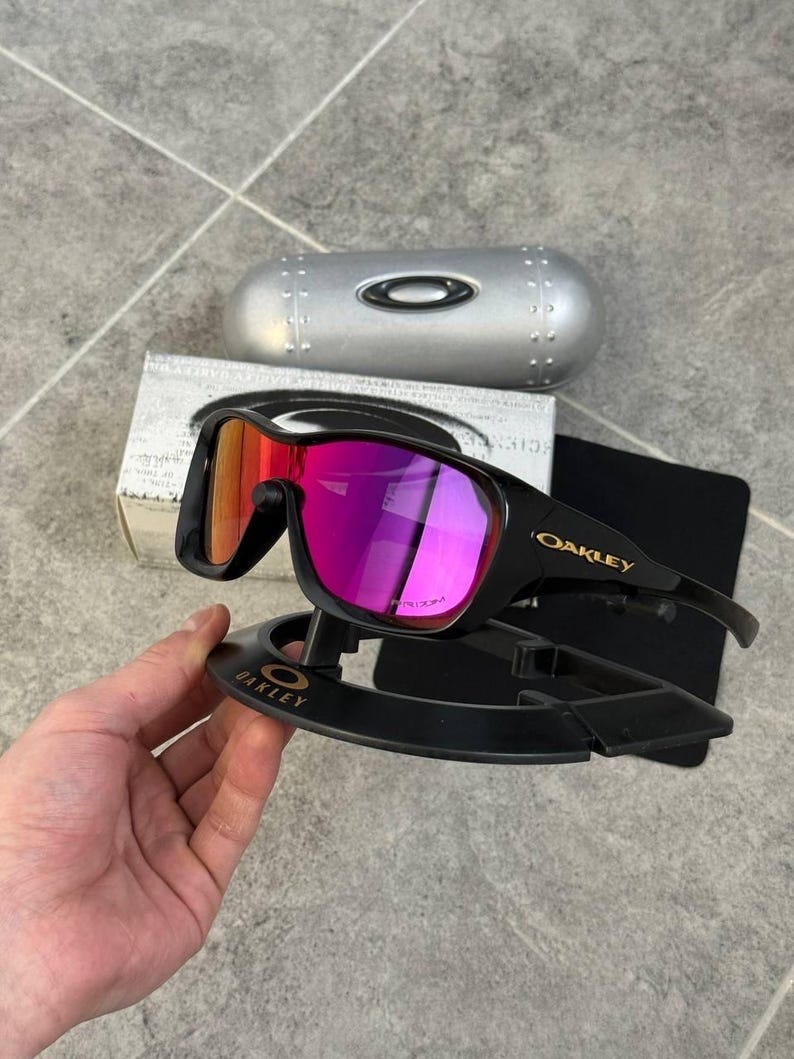 May include: Black Oakley sunglasses with a vibrant pink and orange lens. The sunglasses are displayed on a black stand with the Oakley logo. A silver case and a box are in the background.