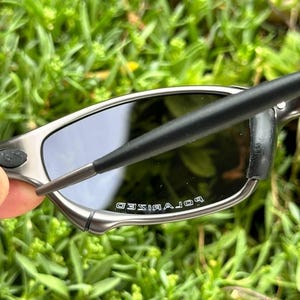 May include: Close-up of polarized sunglasses with a silver frame and dark lenses. The text "POLARIZED" is visible on the lens. The sunglasses are held against a backdrop of green foliage.