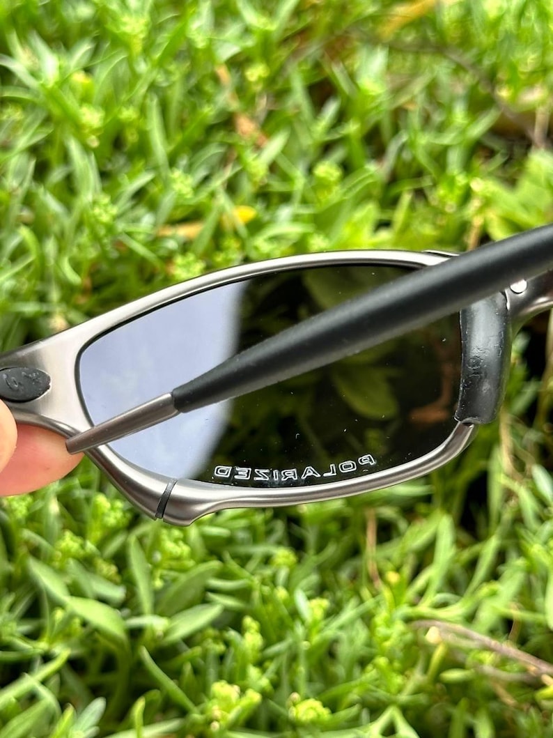 May include: Close-up of polarized sunglasses with a silver frame and dark lenses. The text "POLARIZED" is visible on the lens. The sunglasses are held against a backdrop of green foliage.