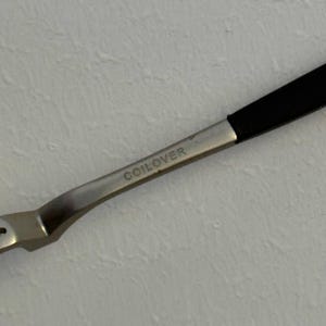 May include: Close-up of a silver metal tool with a black handle. The word "COILOVER" is etched on the metal. The tool is angled, with a curved design.
