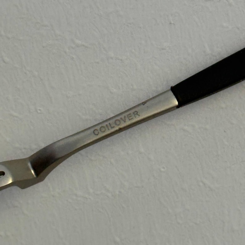 May include: Close-up of a silver metal tool with a black handle. The word "COILOVER" is etched on the metal. The tool is angled, with a curved design.