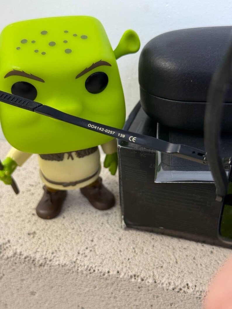 May include: A green Shrek Funko Pop figure with black eyeglasses. The eyeglasses have the text "004142-0257 139 CE" printed on the arm. A black glasses case is in the background.