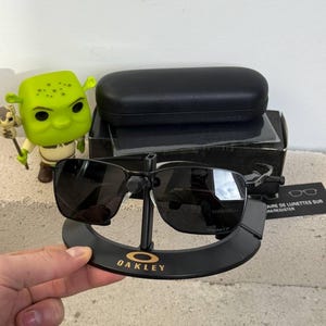 May include: Black Oakley sunglasses with a black case and stand. The sunglasses have a rectangular frame and dark lenses. The stand is black with the Oakley logo in gold. A Shrek figurine is visible in the background.
