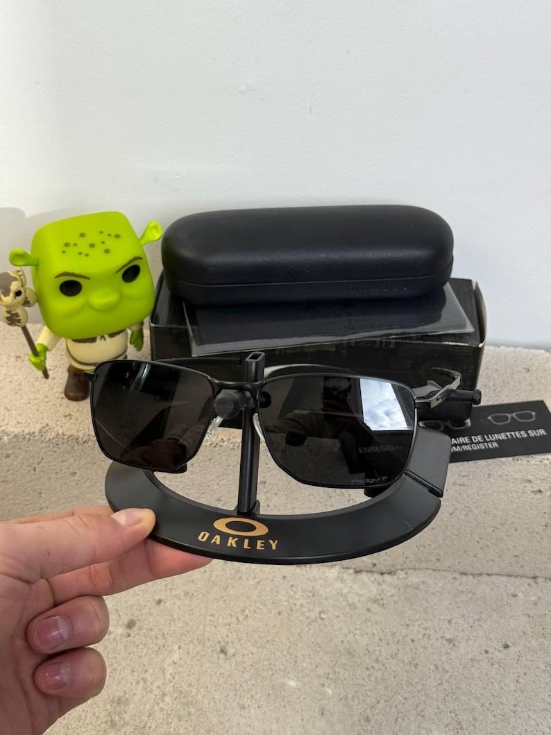 May include: Black Oakley sunglasses with a black case and stand. The sunglasses have a rectangular frame and dark lenses. The stand is black with the Oakley logo in gold. A Shrek figurine is visible in the background.