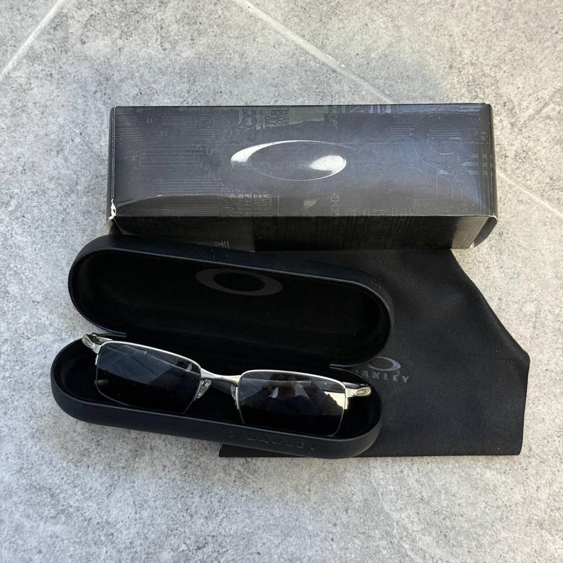 May include: Silver-framed sunglasses with dark lenses are displayed open in a black case, next to a black microfiber cloth and a black box. The case and box have the Oakley logo. The sunglasses have a rectangular shape.