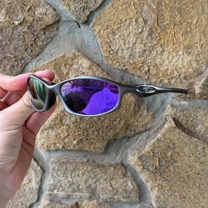 May include: A pair of silver-framed sunglasses with purple mirrored lenses. The sunglasses have a sleek, modern design with a curved frame and dark temples. The lenses reflect the surrounding environment, creating a vibrant purple hue.